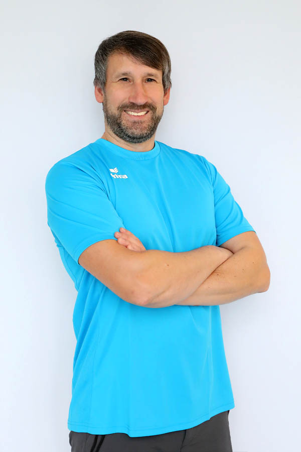 Mann mit blauem T-Shirt und verschränkten Armen lächelt in die Kamera.